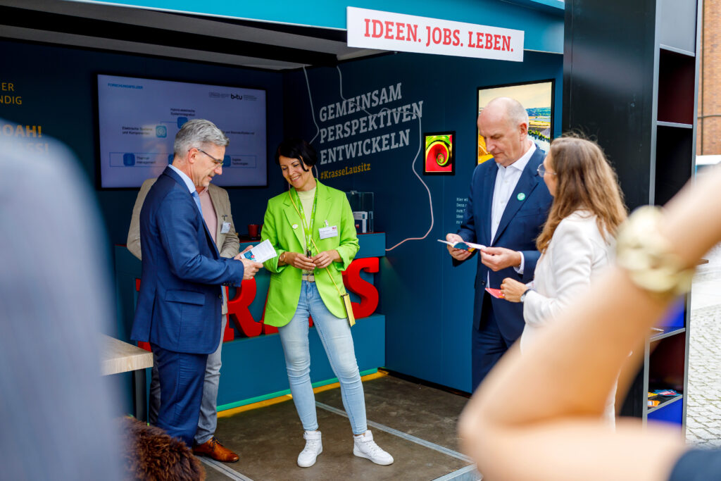 Der Infostand der Imagekampagne hat die Aufschrift: Gemeinsam Perspektiven entwickeln. Auf dem Foto sind verschiedene Menschen zu sehen, darunter Dietmar Woidke, wie sie sich Flyer in de mInfostand durchlesen.