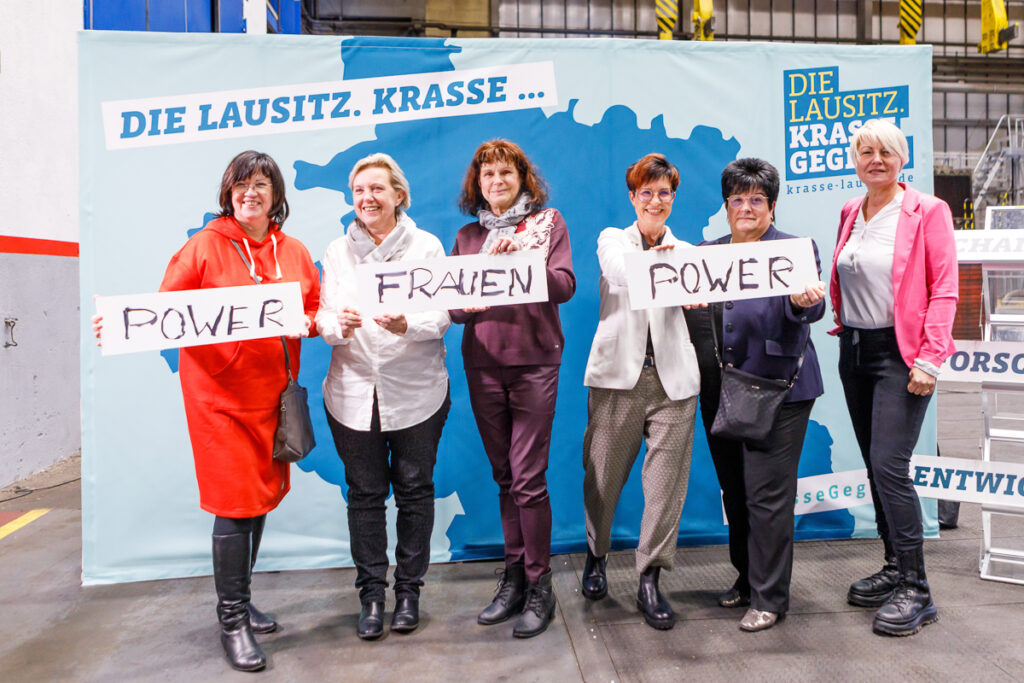 Sechs Frauen stehen vor einer Fotostellwand mit der Aufschrift "Die Lausitz. Krasse Gegend." Dabei halten sie Schilder in der Hand, auf denen die Wörter "Power" und "Frauen" geschrieben steht. 