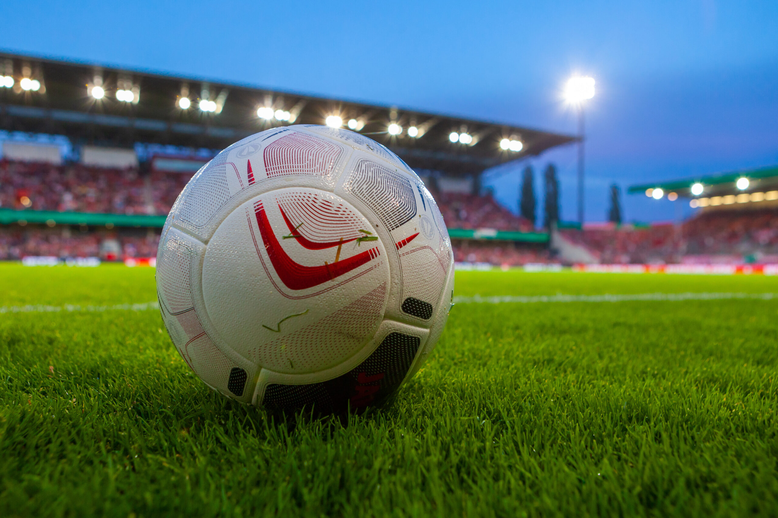 Zu sehen ist ein Fußball auf dem Spielfeld, im Hintergrund eine Tribüne.