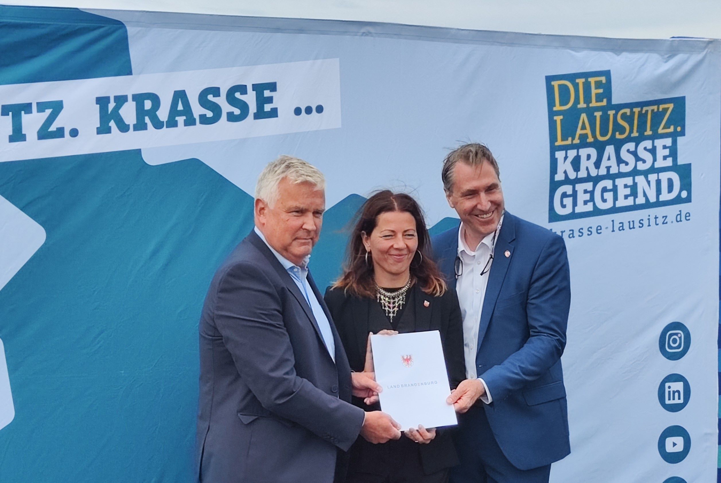Lausitz-Beauftragter Dr. Klaus Freytag, Bürgermeisterin Stadt Cotbus Doreen Mohaupt und Tim Berndt, Geschäftsführer der Entwicklungsgesellschaft Cottbus vor der STellwand Krasse Lausitz halten den Zuwendungsbescheid
