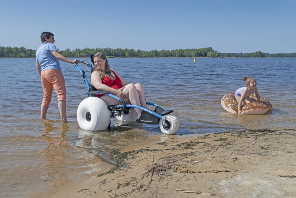 Eine Familie badet an einem See. Im Fokus steht eine junge Frau in einem Amphibien-Rollstuhl