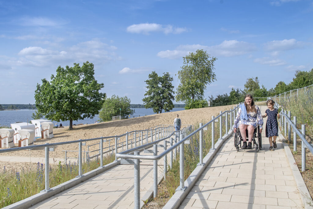 barrierefreier Strandzugang in Form einer Rampe. Im Fokus ist eine Frau im Rollstuhl, wie sie die Rampe nutzt.