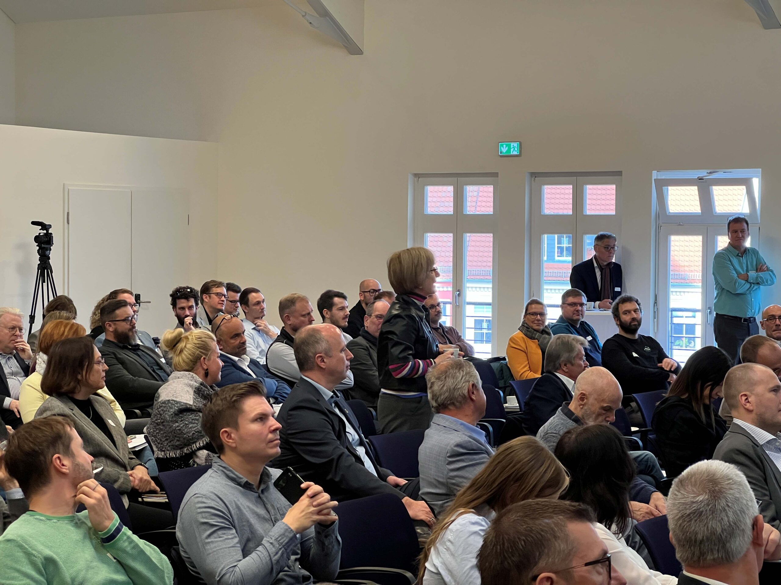 Auditorium in Stuhlreihen. bei der Netzwerkverantaltung. Ein paar Leute stehen im Hintergrund am Fenster und hören zu.