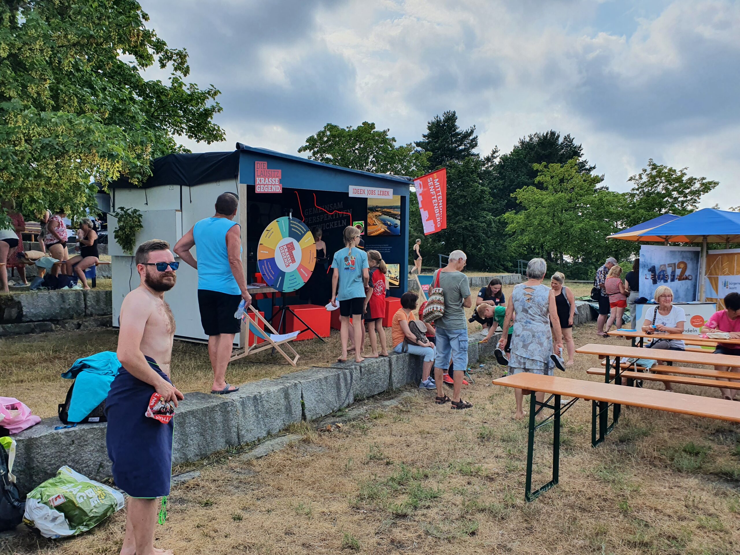 "Krasse Lausitz" Eventstand bei der Eröffnung des Lausitzer Seenlands in Großkoschen. Zu sehen sind Kinder, die vor dem Glücksrad stehen und ein Mann im Vordergrund mit Handtuch um der Hüfte