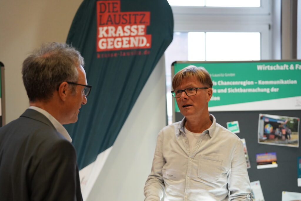 WRL-Mitarbeiter Thomas Berlin im Gespräch am Ausstellungsstand. Im Hintergrund ist eine Beachflag der Krassen Lausitz zu sehen.